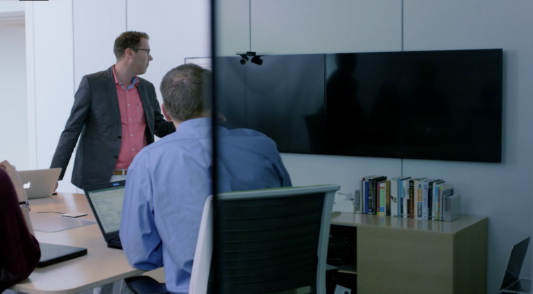 Two men in a conference room looking at a monitor, the image of which is being blocked by casper cloaking film.