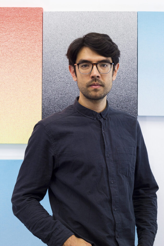 A male artist with glasses posing for a portrait in front of several colorful blocks of color behind him.