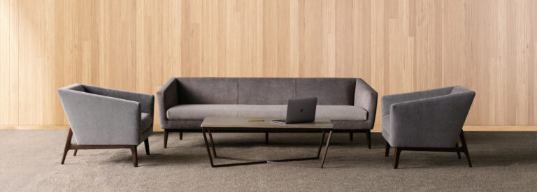 A grey sofa flanked by two side chairs and a coffee table in front of a light wooden wall.