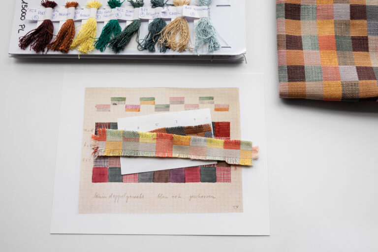 A fragment of a multi-colored checkered textile next to a larger pice of the textile and an assortment of multi color yarn bundles on a white surface.