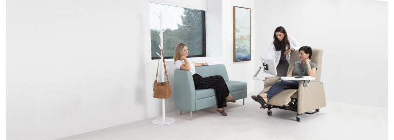 Three women in a healther care office. One, a doctor looking at a table with a women in a patient chair, while another women looks on from a lounge sofa.